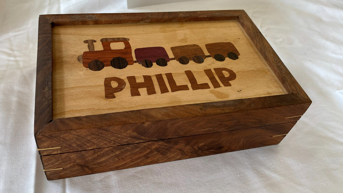 A rectangular wooden box featuring a dark wood frame with mitered corners reinforced by light-colored splines. The lid has a contrasting light wood panel displaying a wood inlay of a train with an engine and three cars in varying shades of reddish-brown and dark wood. Below the train, the name "PHILLIP" is inlaid in large, block-style letters.