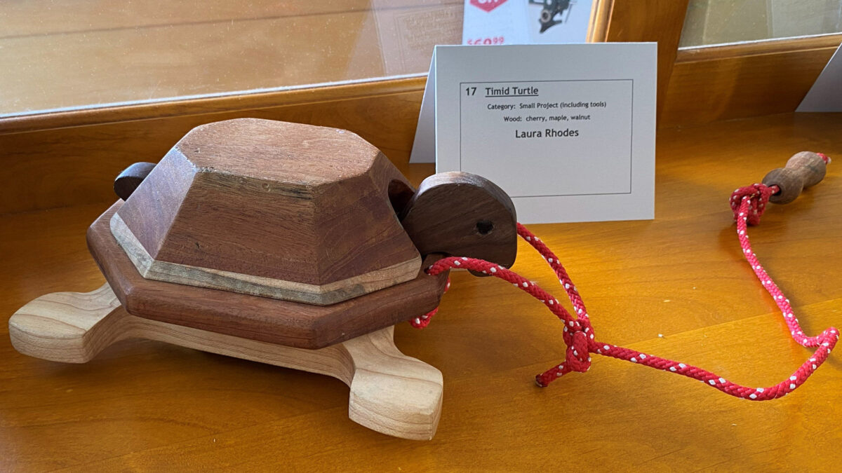A wooden pull-toy turtle featuring a faceted, dark reddish-brown cherry shell layered over a light maple base and legs. The dark walnut head is retracted into the shell. A red-and-white braided rope is attached at the front, ending in a wooden bead. A white display card behind the toy identifies the piece as "Timid Turtle" by Laura Rhodes and notes that it is made of cherry, maple, and walnut.