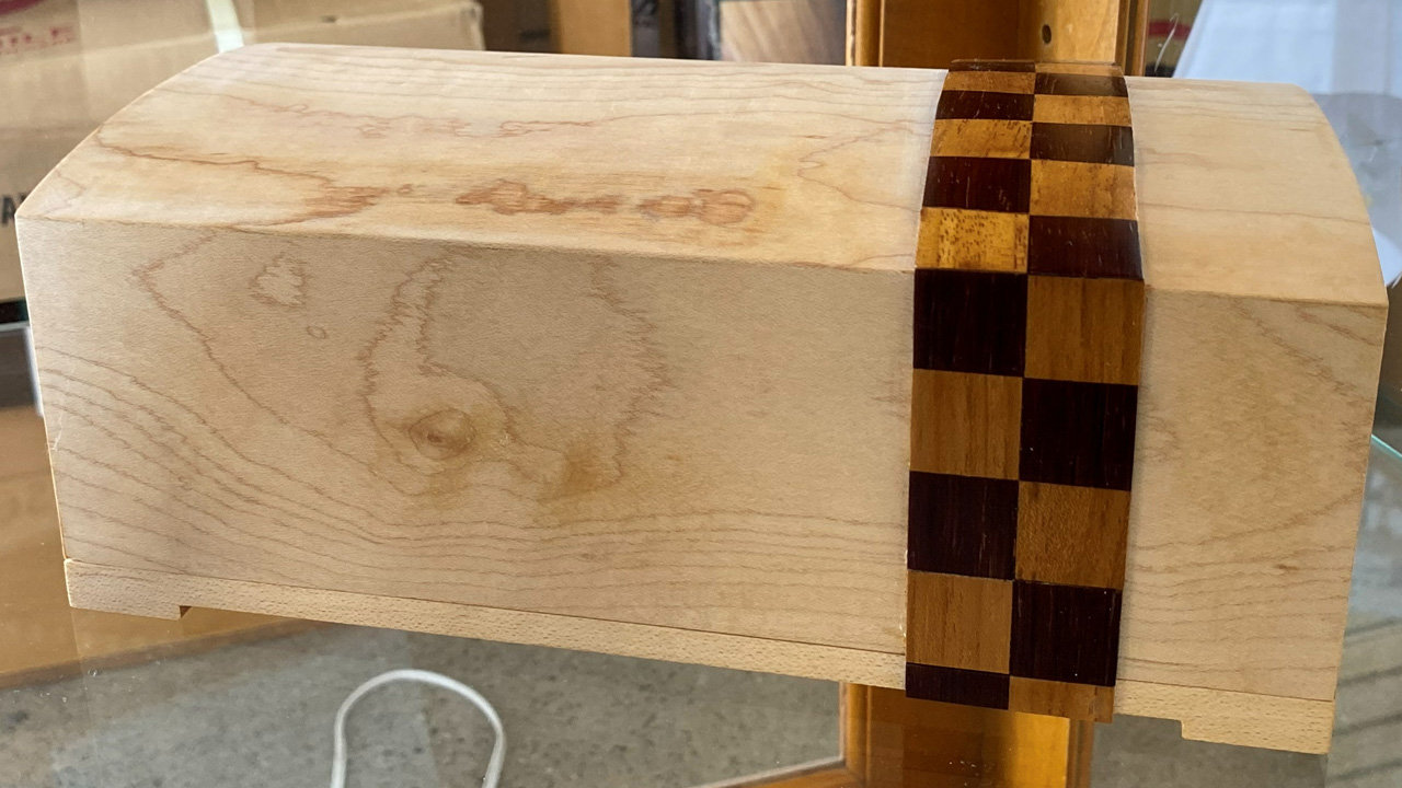 A close-up view of a handcrafted wooden box made from light-colored wood with visible grain. The box features a distinctive vertical inlay, composed of a checkerboard pattern of alternating dark and medium-brown wood squares, designed to resemble a traditional kimono sash or belt.