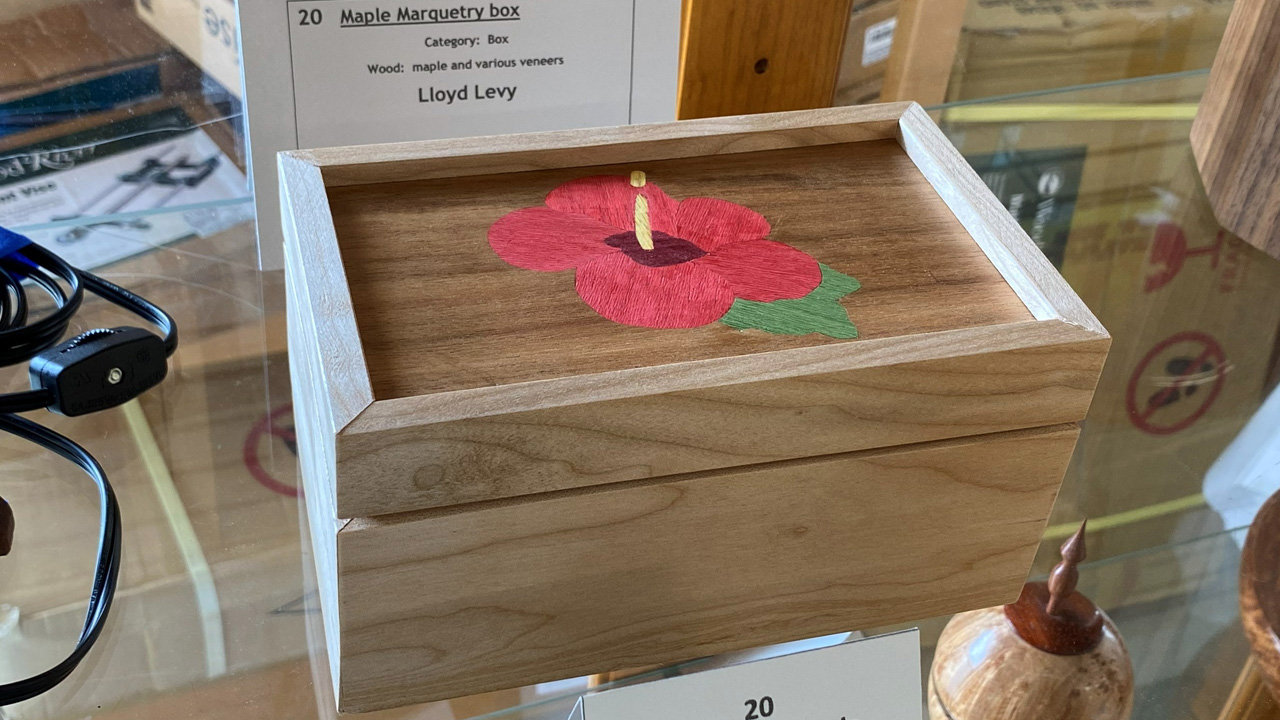 A rectangular wooden box with a light maple frame and darker wood sides. The top of the lid features an intricate marquetry inlay of a red hibiscus flower with a yellow stamen and green leaves. A label identifies the piece as a "Maple Marquetry box" by Lloyd Levy, made of maple and various veneers.