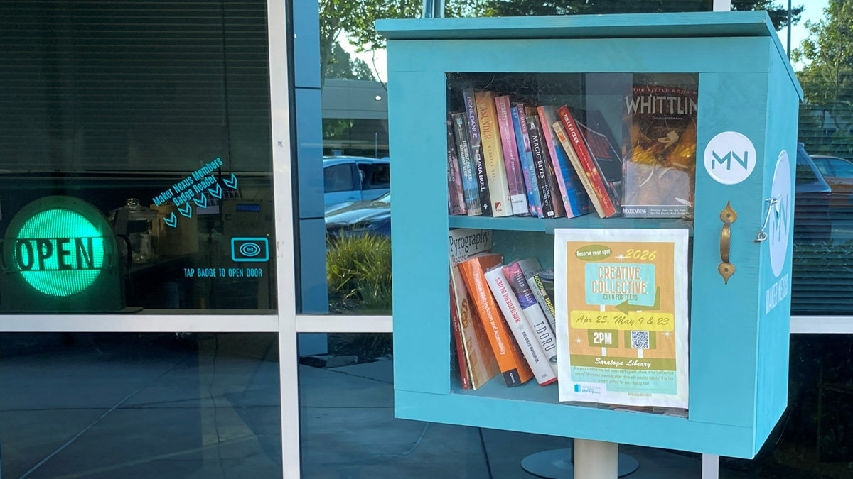 A teal wooden Little Free Library box stands outside the glass entrance of the Maker Nexus building, identifiable by the "OPEN" sign and badge reader instructions on the window, and a circular logo with letters “MN”. The library features a glass-fronted cabinet with two shelves stocked with books; prominently displayed on the top shelf, facing forward, is "The Little Book of Whittling," alongside other craft books. The bottom shelf holds books, including one titled "Pyrography Basics." A flyer for a "Creative Collective Club for Kids 2026" event is taped to the glass door.