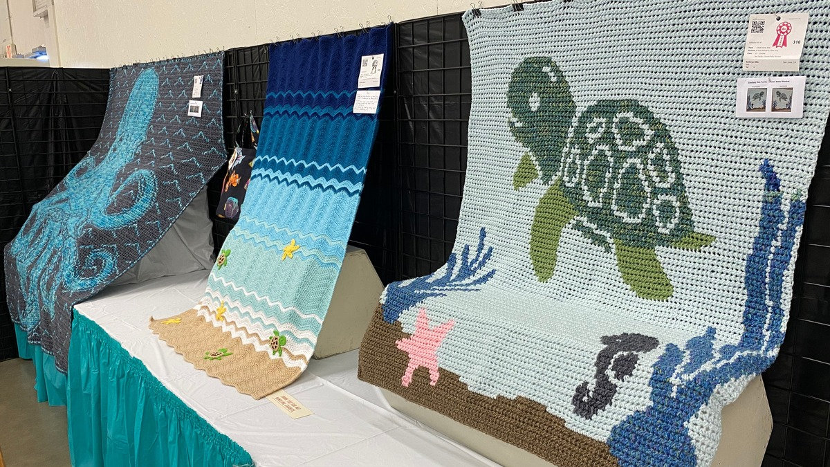 Three marine-themed crochet blankets featuring a sea turtle, octopus, and ocean waves are on display at the Santa Clara County Fair in San Jose, California. A red prize ribbon is attached to the turtle blanket.