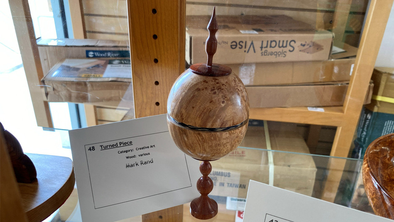 A turned wooden piece displayed on a glass shelf, identified by a white label as "Turned Piece" by artist Mark Rand. The project is categorized as "Creative Art" and made of "various" woods. The object features a large spherical body crafted from light-colored wood with a visible grain pattern, separated by a thin dark band. It sits atop a turned pedestal base of darker reddish-brown wood and is capped with a matching pointed finial.