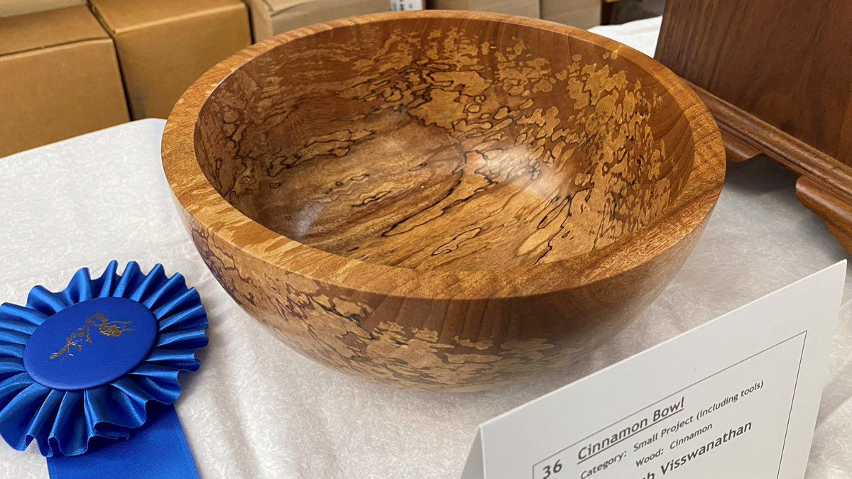 A polished wooden bowl with a striking, intricate grain pattern of swirling golden-browns and dark streaks sits on a white tablecloth. To the left lies a blue rosette prize ribbon. A white identification card in the foreground labels the piece as the "Cinnamon Bowl," made from Cinnamon wood by an artist named Mayuresh Visswanathan.