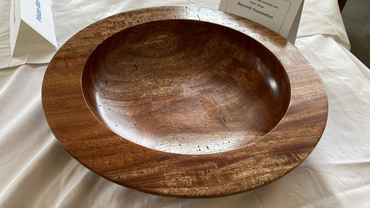 A large, shallow wooden bowl turned from Kamani wood. It features a wide, flat rim and a smooth, concave interior with a high-gloss finish. The wood displays a rich, warm brown tone with prominent, swirling grain patterns across the surface. Mayuresh Visswanathan created the piece.