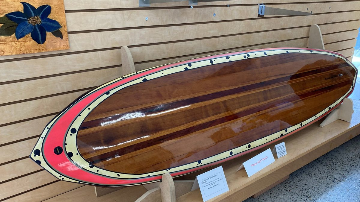 A handcrafted wooden surfboard, styled as a classic Malibu longboard, resting on wooden display stands at the woodworking show organized by Bay Area Woodworkers Association (BAWA). The board features a high-gloss finish and is constructed from laminated strips of wood in varying shades of reddish-brown and golden-brown, creating a striped pattern down the center. The edges are decorated with a cream-colored border featuring black polka dots and a thick, curved red band near the nose. Black lines separate the decorative bands from the wood veneer. Small white information cards are visible on the shelf in front of the board.