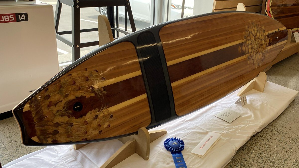 A custom wooden surfboard displayed on wooden stands at the woodworking show at Woodcraft San Carlos. The board is constructed using a strip-building technique, featuring alternating longitudinal stripes of light amber, medium brown, and a central dark wood stripe (likely walnut). The surface is heavily decorated with large inlays of dried flowers and botanical specimens encased in clear epoxy resin. One large floral section is at the tail (left side), where a black leash plug is visible, and another is located further up the deck. A dark, possibly carbon fiber or black wood, transverse band crosses the board near the middle. The maker's signature is visible in the resin near the nose on the right. A blue award ribbon rests on the white cloth in the foreground.