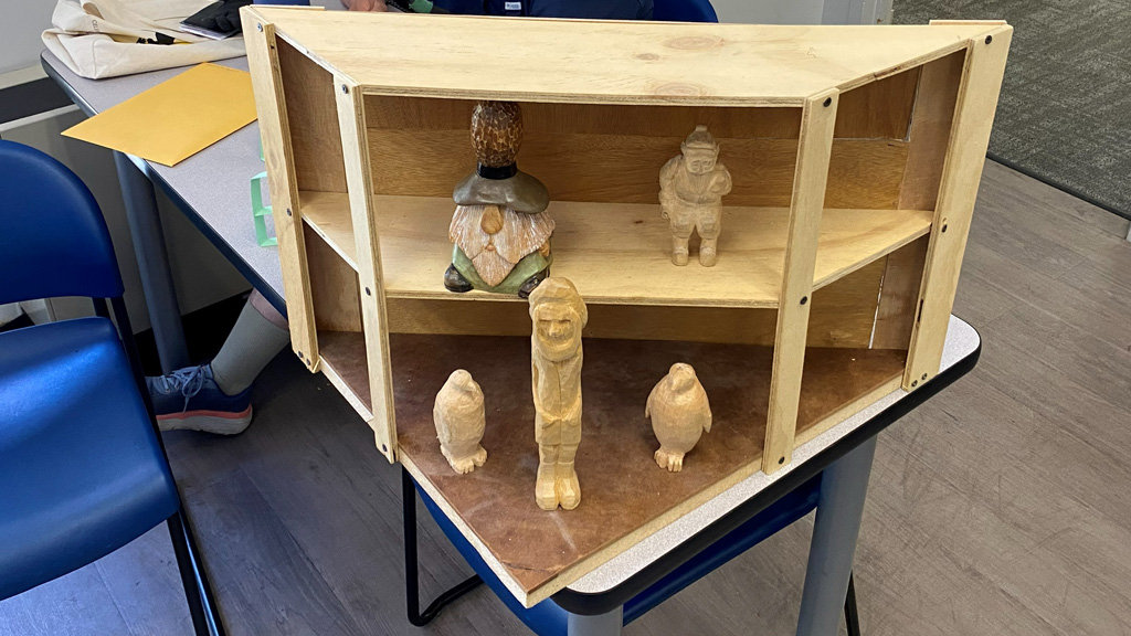 A light-colored wooden corner shelving unit, which appears to be an early prototype made of plywood with visible screws, sits on a table displaying several hand-carved wooden figures. On the upper shelf, there is a painted figure resembling a gnome alongside a smaller standing human figure. The bottom surface of the case features three unpainted carvings: two small penguins flanking a taller, slender human figure in the center.