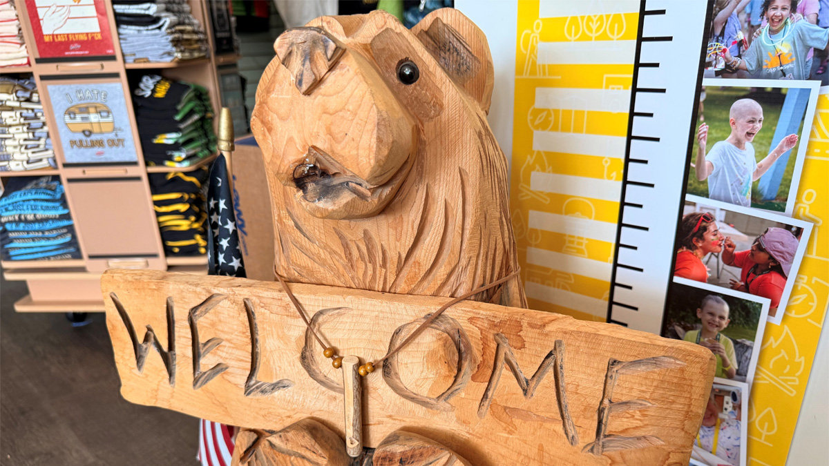 A rustic wooden carving of a bear holding a wooden sign that reads "WELCOME" in deeply carved letters. The bear has a black bead for an eye, charred detailing around its snout and mouth, and wears a necklace made of twine and small wooden beads. In the background, there is a display rack of folded T-shirts on the left and a yellow poster board featuring photos of smiling children on the right, suggesting this carving serves as a welcome figure at a camp.
