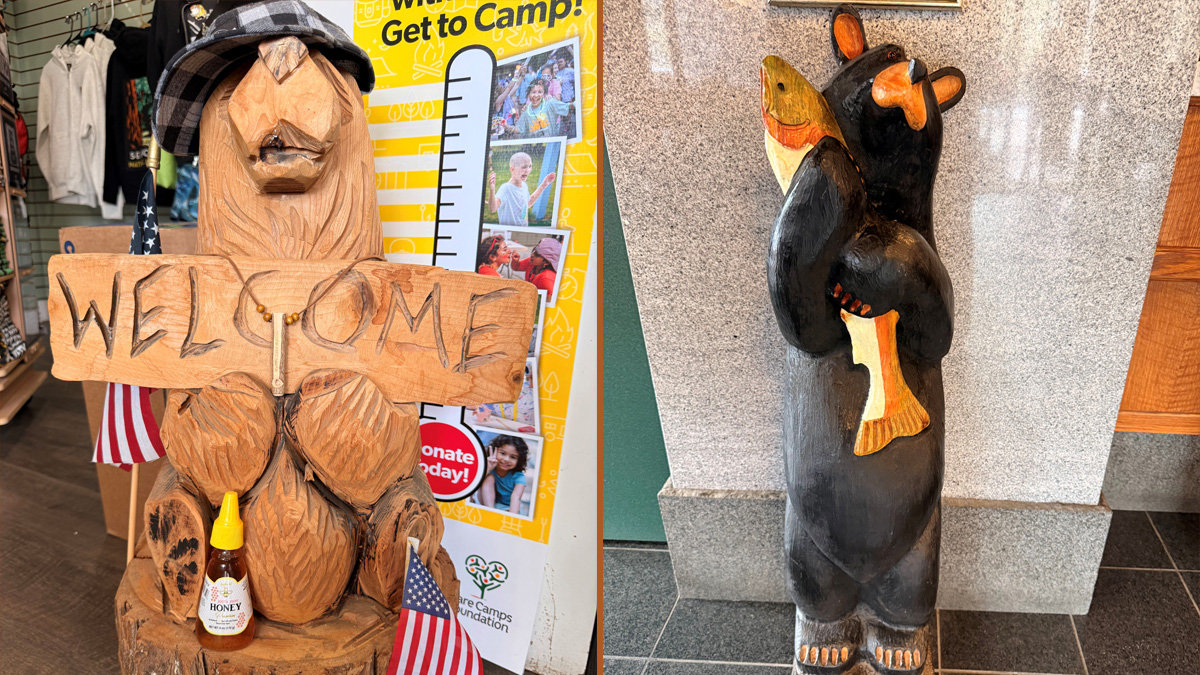Two side-by-side photographs of hand-carved wooden figures. On the left, a rustic light-wood carving of an animal with large buck teeth and a plaid hat holds a "WELCOME" sign, with a bottle of honey and small American flags at its base. On the right, a painted black bear stands upright on a tiled floor, clutching a yellow fish against a speckled stone column.