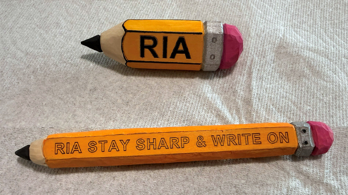 Two hand-carved orange wooden pencils lying on a white textured cloth. The top pencil is short with the name "RIA" painted in black. The longer bottom pencil features the engraved message "RIA STAY SHARP & WRITE ON." Both feature realistic details including sharpened black tips, silver ferrules, and pink erasers. The pattern for the carvings was created by the pencil-shaped name badge generated at READ N TRY website, https://readntry.com/woodcarving-pattern-generator-pencil-name-badge/