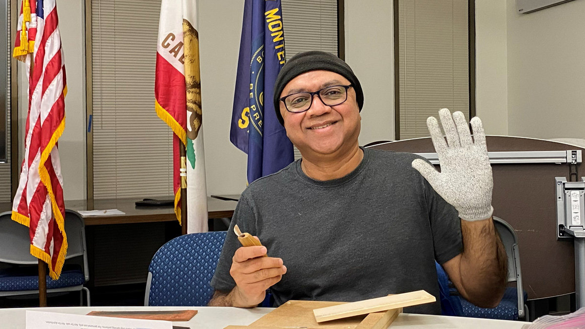 A smiling man wearing a black beanie, glasses, and a grey T-shirt sits at a table covered with pieces of light-colored wood (basswood). He holds a small chip-carving knife in his right hand and raises his left hand to show off a grey protective glove. Behind him stand three flags: the United States flag, the California state flag, and a blue organizational flag of Silicon Valley Monterey Bay Council of Scouting America.