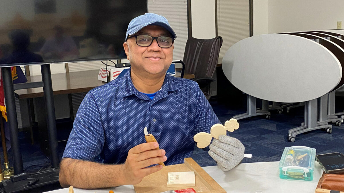 A smiling man, Nitin, is wearing a blue cap and glasses, whittling a basswood heart-shaped ornament with a small carving knife.