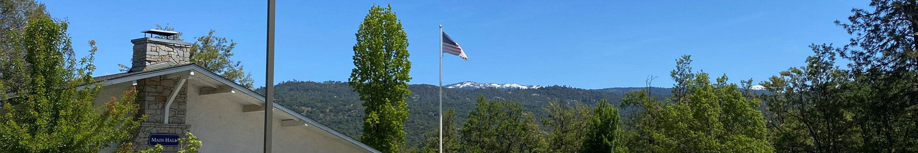 A wide panoramic shot taken on a sunny day shows a scenic view from the Oakhurst Community Center featuring the corner of a beige stucco building with a prominent stone chimney and a blue sign reading "MAIN HALL." An American flag flies from a tall white pole near the center amidst lush green trees, while the background reveals a sweeping horizon of forested hills leading up to distant mountain peaks dusted with snow under a clear blue sky.