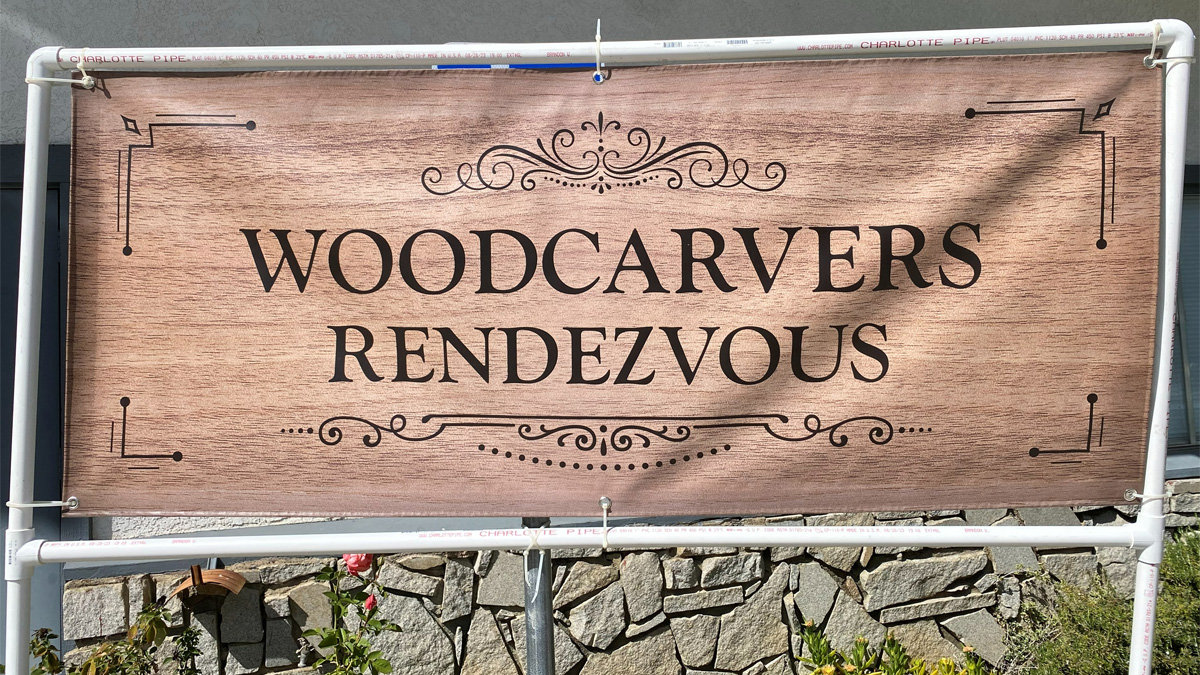 A large rectangular banner with a light brown, faux wood-grain texture hangs from a white PVC pipe frame outdoors. The words "WOODCARVERS" and "RENDEZVOUS" are printed in bold black serif letters across the center, flanked by ornate scrollwork and corner decorations. Behind the sign, an irregular grey stone wall and some green plants with red flowers are visible at the bottom.