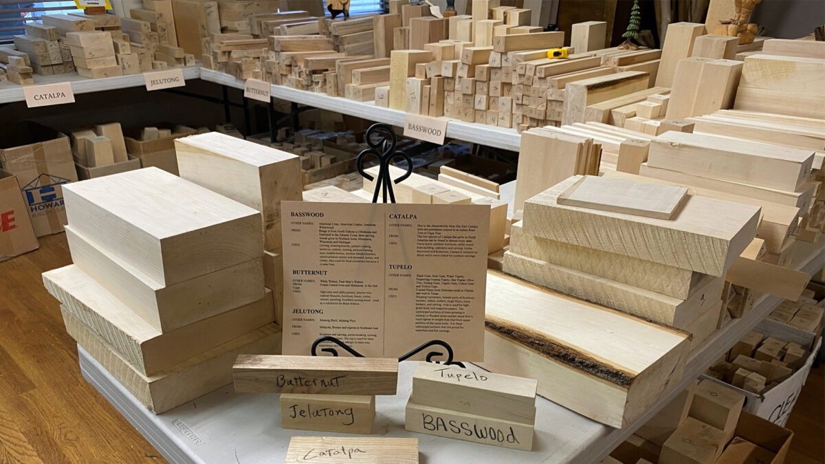 This image displays a supplier's table at the Oakhurst Woodcarvers Rendezvous, densely packed with various types of lumber intended for woodcarving. In the foreground, small sample blocks labeled by hand ('Butternut,' 'Jelutong,' 'Tupelo,' 'Basswood,' and 'Catalpa') are arranged around an informational card describing each wood type's origin and uses. Behind these samples are large stacks of raw, rectangular lumber blocks in shades of cream and pale tan, with background shelves stretching across the frame, stacked high with hundreds more pieces of wood organized by type under hanging tags.