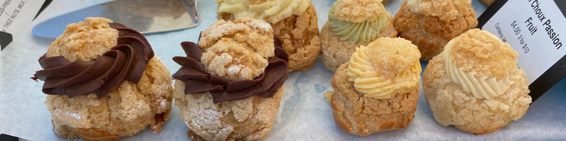 Close-up of assorted cream-filled choux pastries with crumble tops on parchment paper at a San Jose farmers market, serving as a reference for a woodcarving workshop.