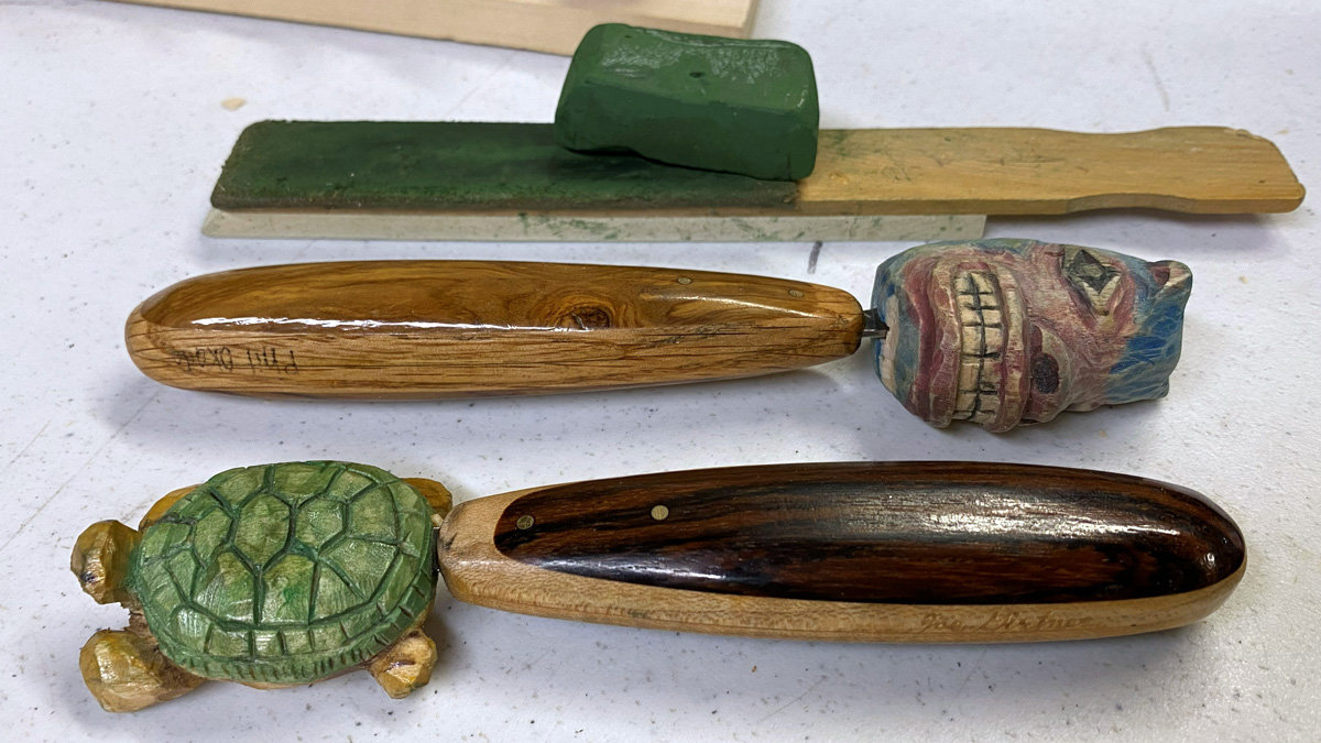 Three handmade woodworking items are arranged vertically on a white surface: a DIY sharpening strop made from a paint stir stick and leather with a block of green compound placed on top, a carving knife with a colorful tiki-style blade cover featuring prominent teeth, and another knife whose blade is hidden inside a detailed green turtle figurine. Phil Okada used these tools at the 2026 Oakhurst Woodcarvers Rendezvous.