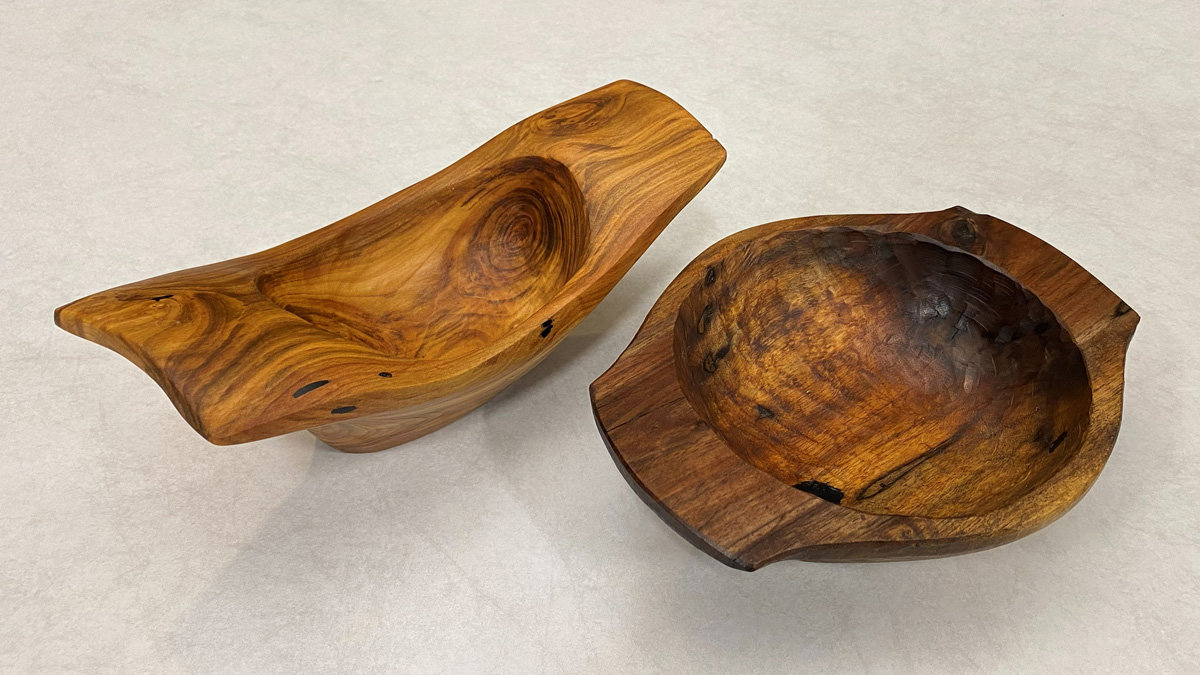 Two hand-carved wooden vessels by Pranay Agrawal are displayed side by side on a neutral, light gray background. On the left is an elongated, canoe-shaped bowl crafted from light honey-colored wood with distinct swirling grain patterns and small dark knots. To its right sits a deeper, rounder bowl with handles and a flat rim; it features a contrasting dark brown textured interior. The image is featured in the SCVC woodcarving newsletter.