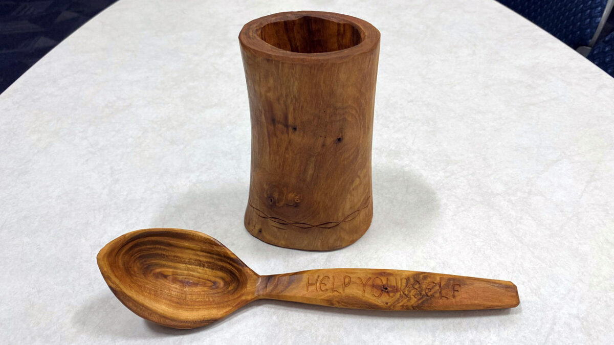 A wooden shrink box standing upright next to a large wooden spoon lying horizontally on a white surface. The cylindrical box showcases rich wood grain and features decorative twisting lines carved near its base. The spoon has the phrase "HELP YOURSELF" carved into its flat handle, highlighting the craftsmanship of the woodworking project.