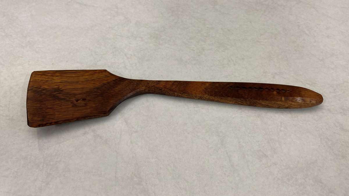 A hand-carved wooden spatula with a wide head and long handle lying on a light surface.