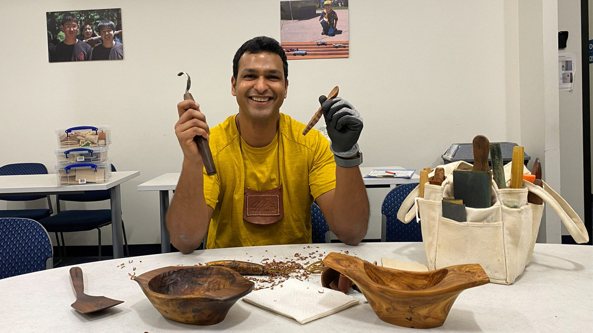 Pranay Agrawal smiles broadly while seated at a table covered in wood shavings during a woodcarving session for the Santa Clara Valley Carvers. Wearing a yellow shirt and a small leather apron, he holds up a curved carving tool (hook knife) in his right hand and a roughed-out wooden spoon handle in his gloved left hand. The foreground features two carved wooden bowls and a spatula, while a canvas tote bag packed with various chisels and gouges sits to the right.