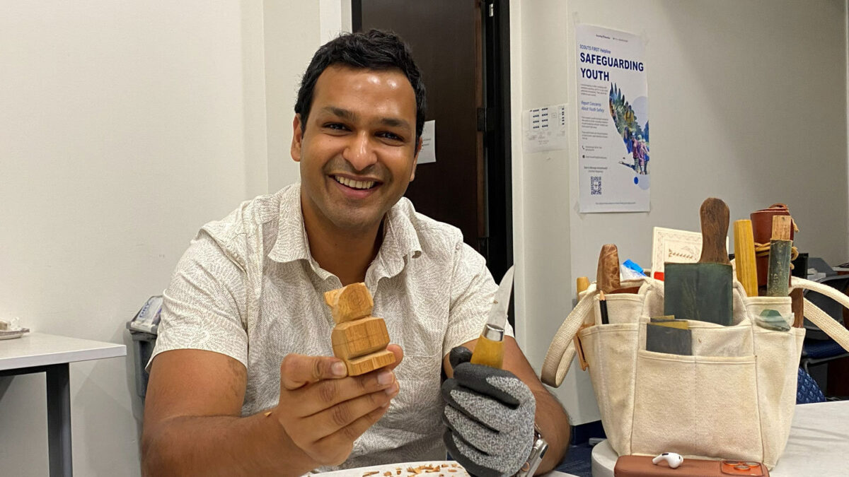 Pranay Agrawal smiles while holding a small wooden carving and carving knife, wearing a protective glove at a Santa Clara Valley Carvers meeting in San Jose, California.