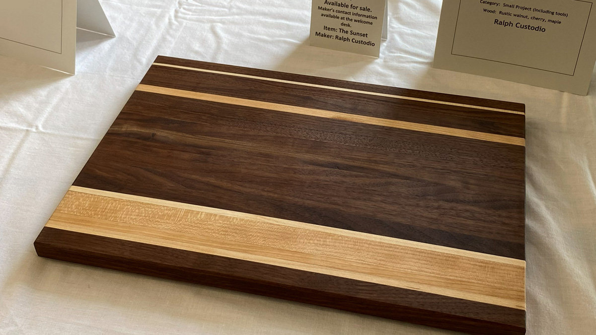 Rectangular wooden cutting board by Ralph Custodio featuring a striped design. The board has a large central panel of dark, richly grained wood (walnut) accented by a thin, light-colored strip near the top edge and a wide band of light wood (maple and cherry) across the bottom section. The wood has a smooth matte finish.