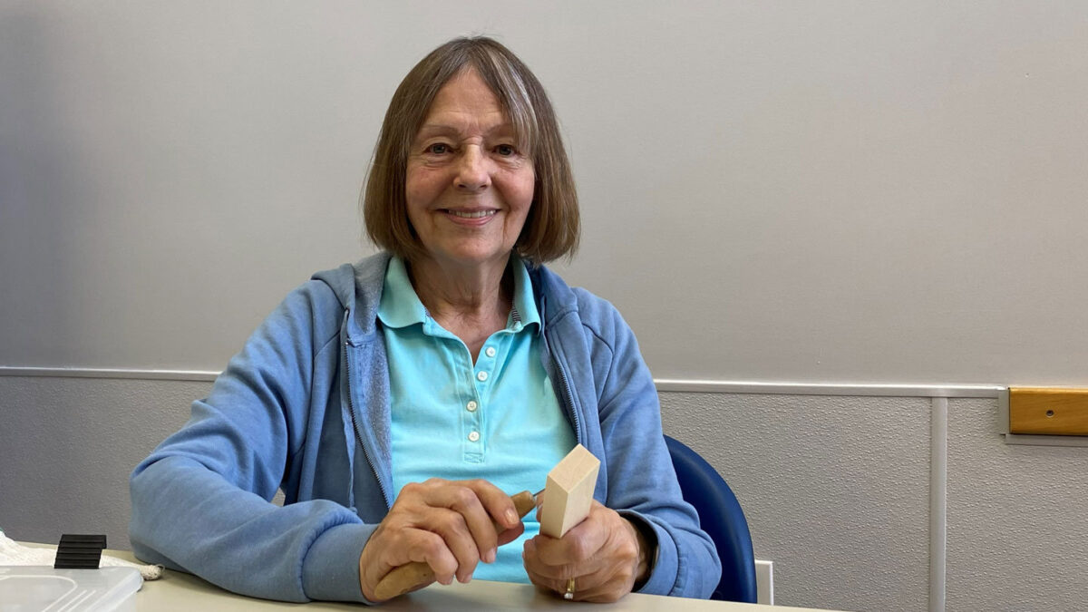 Rena Gooding smiles while holding a light-colored wooden block (basswood) and a carving knife during a Santa Clara Valley Carvers meeting in Los Gatos, California.