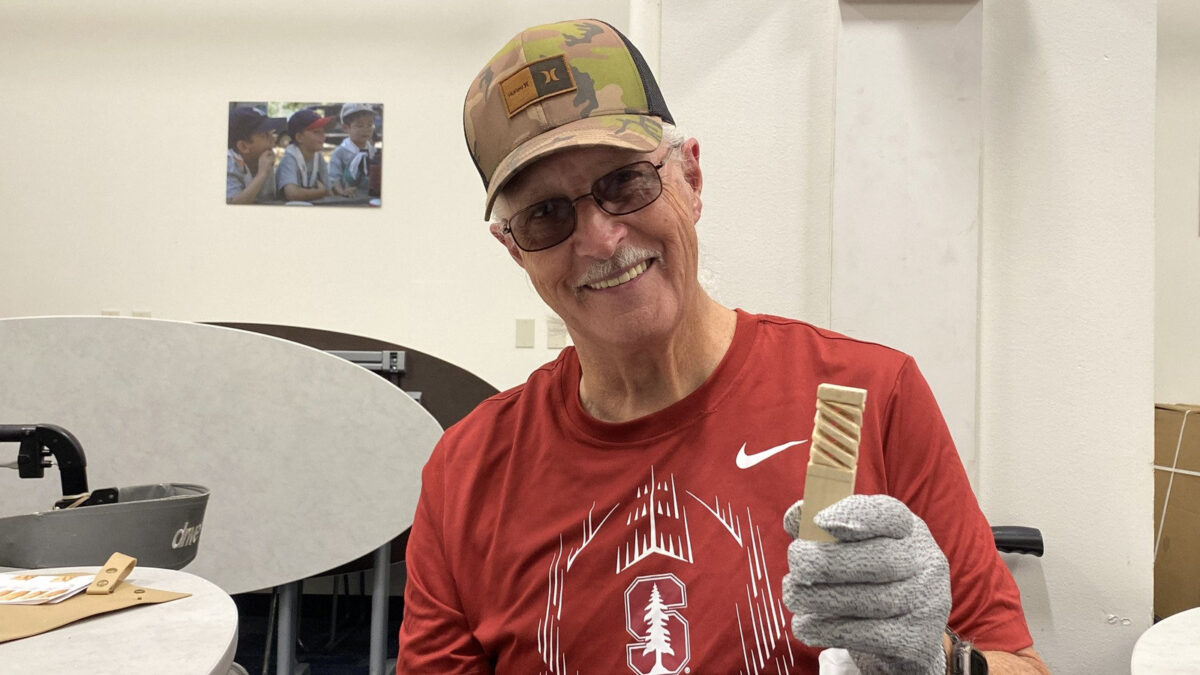 Roger Williams, wearing a red Stanford T-shirt and camo hat, displays a small basswood block with a carved spiral pattern at a Santa Clara Valley Carvers meeting in San Jose, CA.