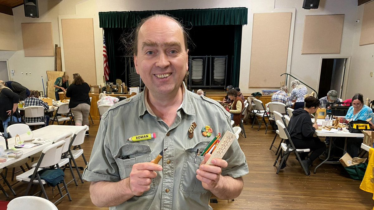 A smiling man, identified by his pencil-shaped nametag as Roman, stands in the foreground holding a woodcarving knife and a small wooden block featuring a chip-carved ornament. He wears a grey button-down shirt adorned with pins. The background shows a busy community hall during the Oakhurst Woodcarvers Rendezvous in 2026, with participants seated at white tables, working on various projects near a stage with green curtains.