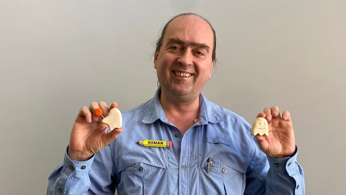 Roman Chernikov smiles while displaying two small wooden Halloween ghost carvings, one holding a tiny pumpkin, at a Santa Clara Valley Carvers meeting in Los Gatos, California.