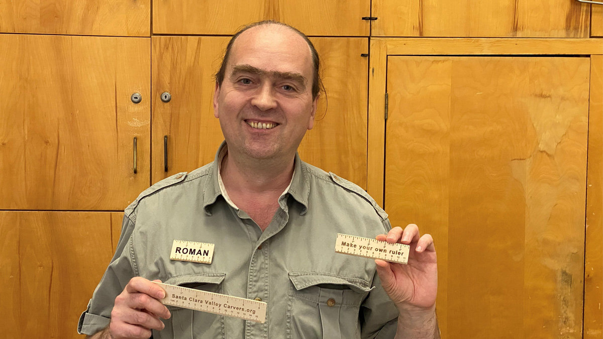 Roman Chernikov smiles at the camera while standing in front of light wood cabinetry, holding up two custom-made wooden rulers. He wears a green utility shirt with a name tag reading "ROMAN" on his chest. In his right hand (viewer's left), he holds a ruler featuring measurement scales and the text "Santa Clara Valley Carvers.org," while in his left hand (viewer's right), he holds a smaller wooden ruler engraved with the phrase "Make your own ruler."