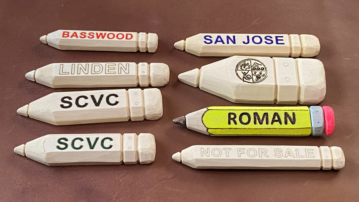 A collection of hand-carved basswood name badges shaped like pencils, each featuring a tapered point and a flat eraser end. The badges display various text in different colors, including "BASSWOOD" in red and "SAN JOSE" in blue, while one is painted yellow with the name "ROMAN" and colorful ends. All were whittled from single pieces of wood using only one carving knife.