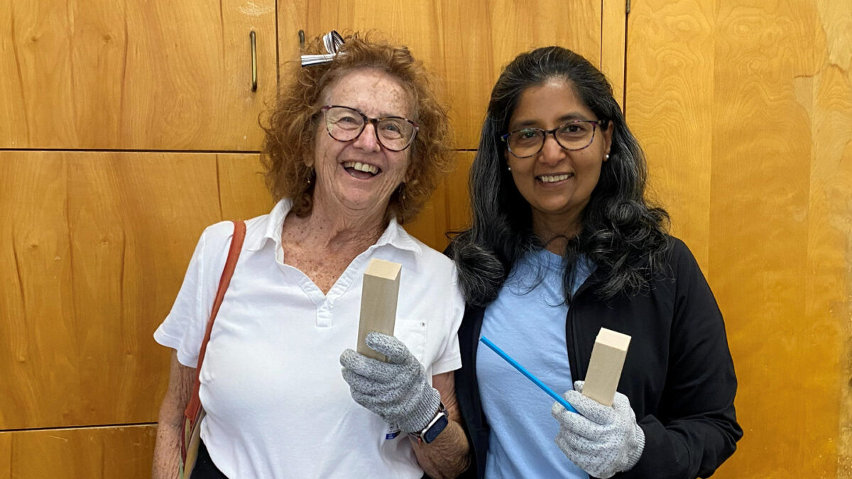 Members Sally Barlow-Perez (left) and Padmaja Nandigam (right) smile while holding blocks of basswood and wearing protective gloves.