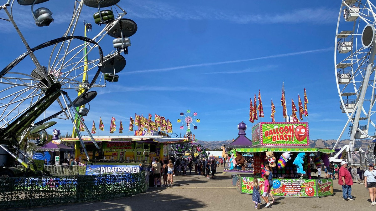Sunny midway scene at the Santa Clara County Fair in San Jose, featuring a Ferris wheel, "Balloon Blast" game booth, and visitors strolling under a blue sky.