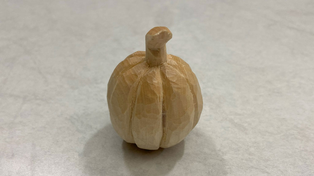 A small pumpkin with a curved stem carved from basswood by Sarah Liu. It is 1.5 inches in diameter.