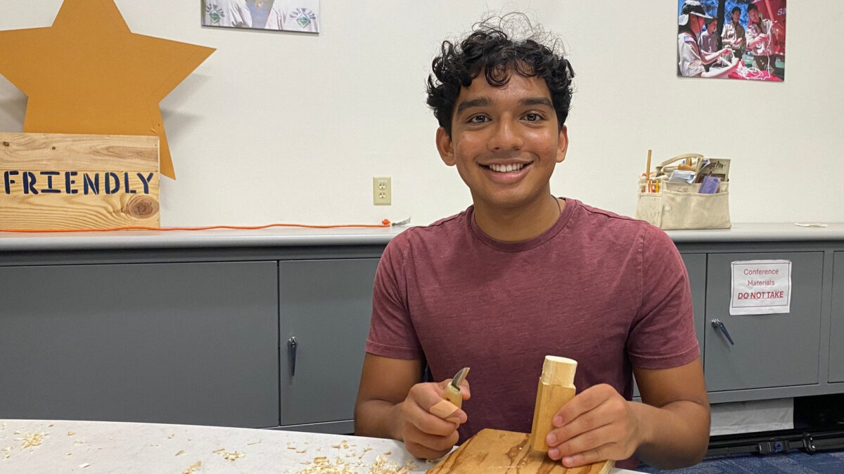 Sarvajith Kujuluva smiles while holding a carving knife and wood block at a Santa Clara Valley Carvers meeting in San Jose, California, surrounded by wood shavings.