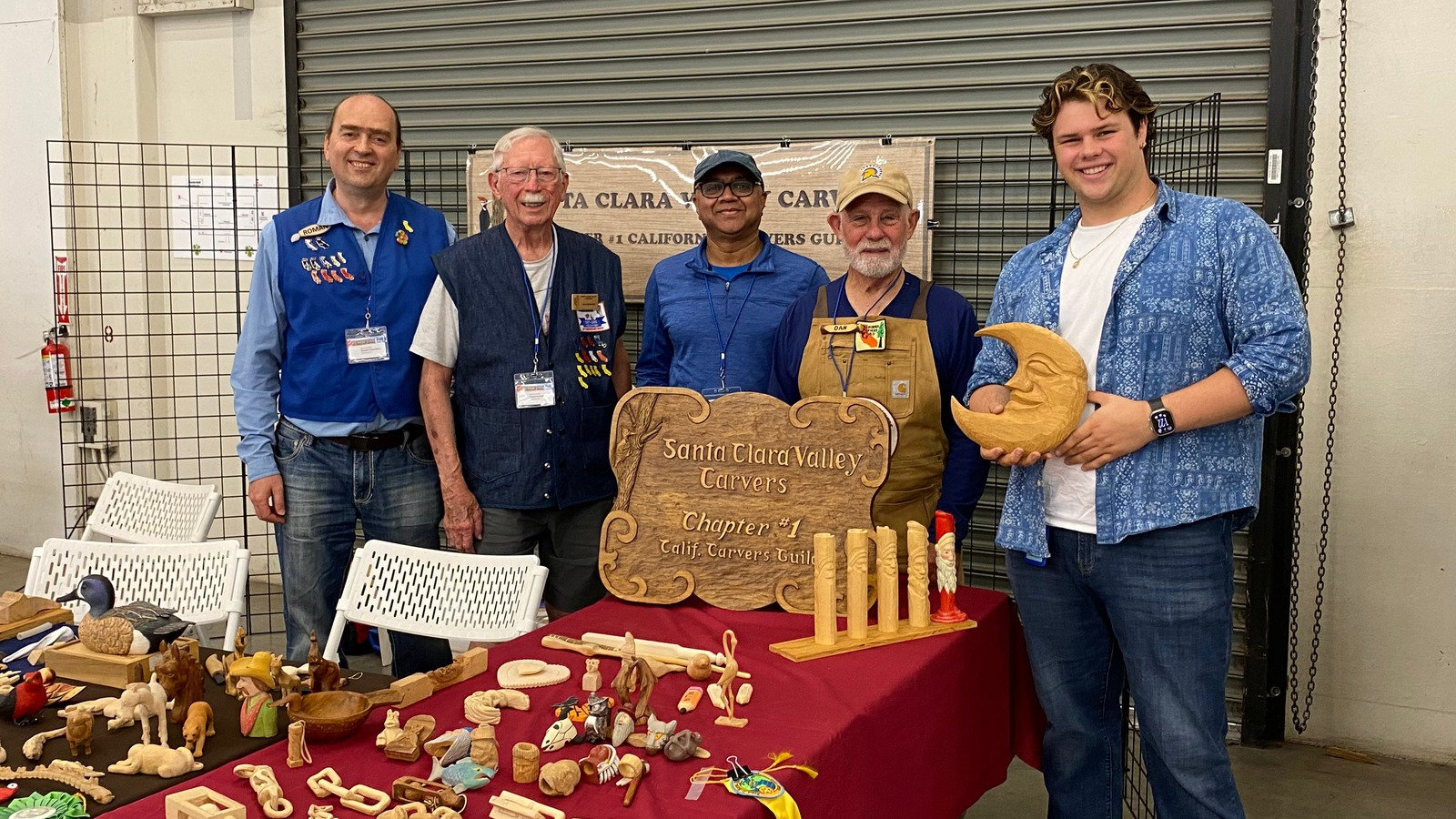 Members of the Santa Clara Valley Carvers and Shea De'Arman pose behind a table of woodcarvings at the 2026 Santa Clara County Fair, featuring a carved club sign and wooden moon.