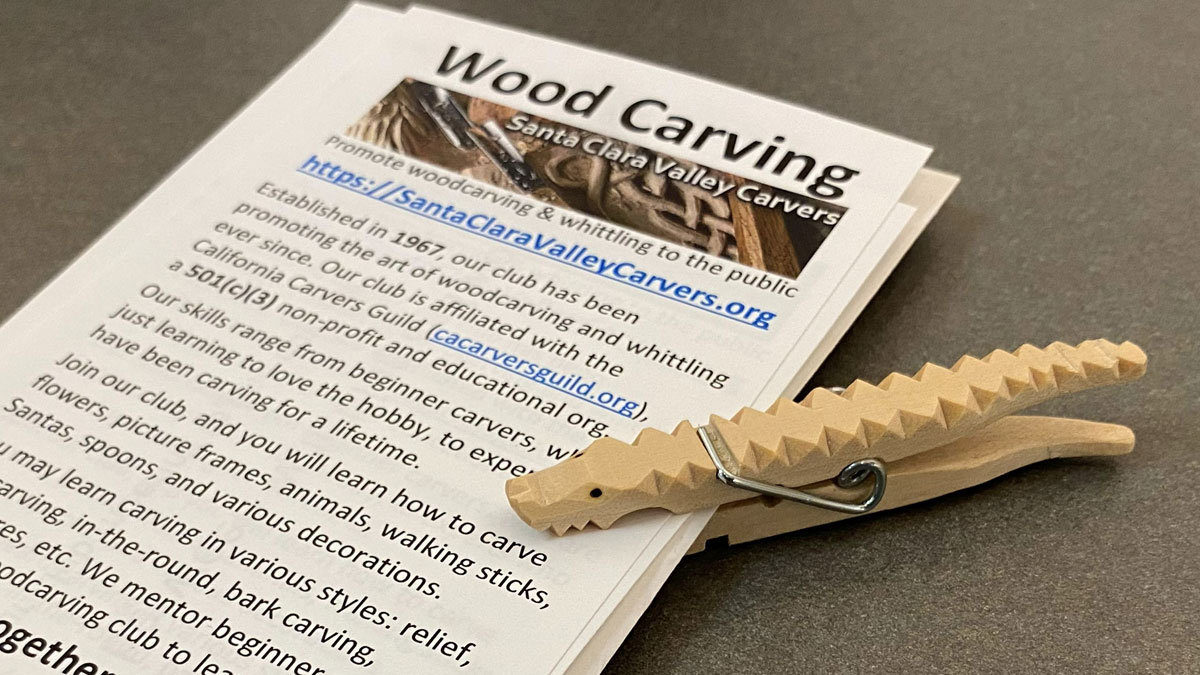 A stack of woodcarving club's flyers held together with a clothing pin carved to resemble an alligator.