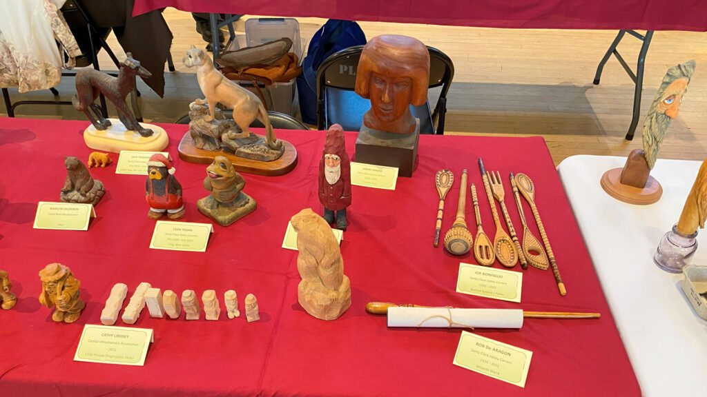 A table full of woodcarvings from several artists: carved animals, spoons, and caricatures.