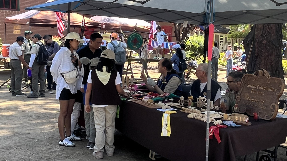 At the Scout-O-Rama event at History Park, members of the Santa Clara Valley Carvers demonstrate the craft of woodcarving at an outdoor booth. Three men (Roman Chernikov, David Osterlund, and Mike Campi) sit behind a table covered in a brown cloth, which is filled with various small wooden carvings, figurines, and tools. One carver, Roman, gestures with his hand while talking to a woman in a white shirt and hat, and a child wearing a brown scout vest. A carved wooden sign on the right side of the table reads "Santa Clara Valley Carvers Chapter 1, California Carvers Guild." In the background, other tents, flags, and attendees are visible under sunny skies.