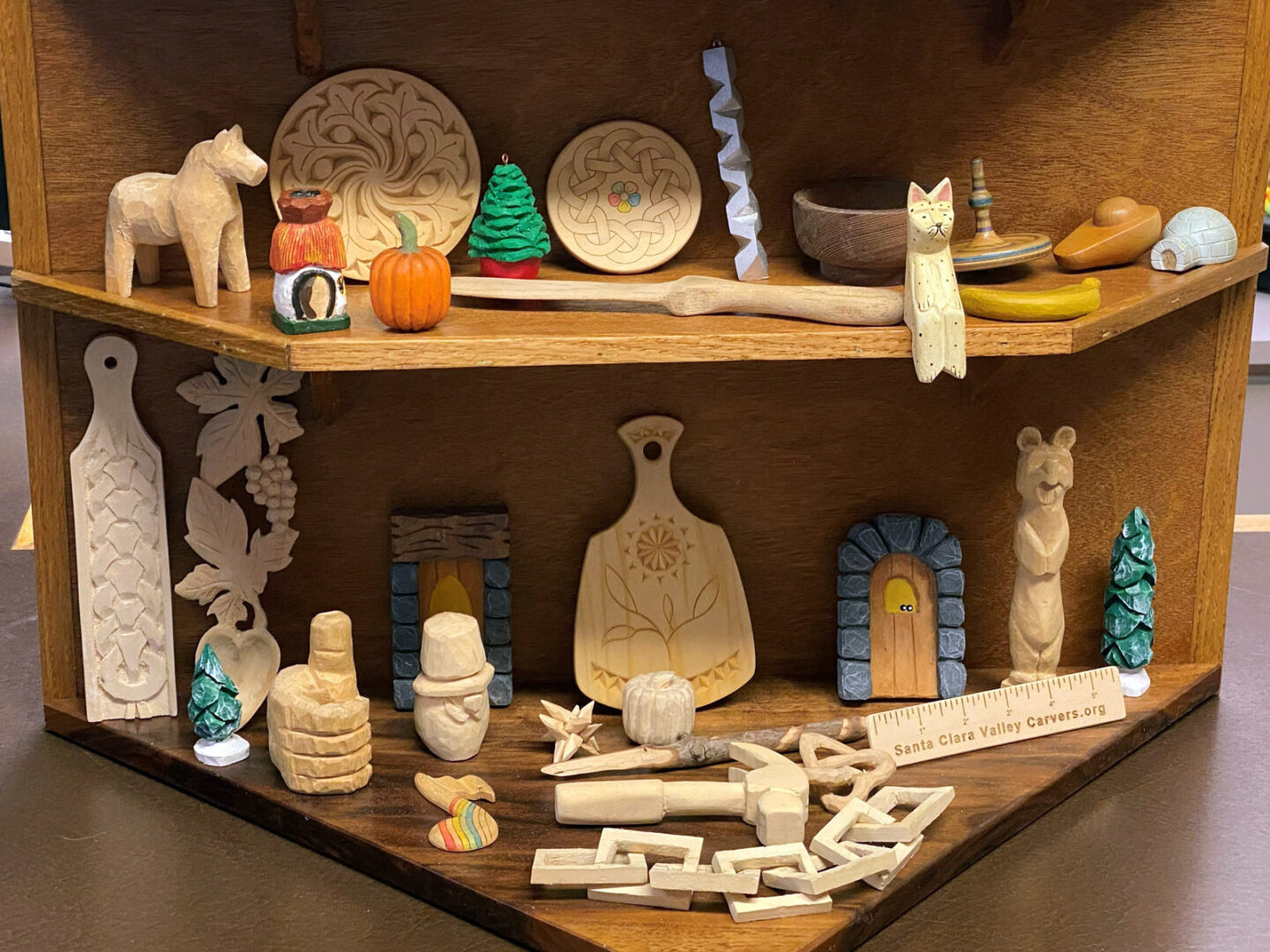 A collection of hand-carved wooden projects displayed on a two-tiered wooden corner shelf unit. The top shelf holds a variety of items, including a stylized horse, small painted figurines like a mushroom house and pumpkin, decorative wall plaques with Celtic and swirl designs, a green tree, a wooden bowl, a standing cat figure, and a long wooden spoon resting across the shelf. The bottom shelf features functional and decorative pieces such as a tall DIY decorated strop with braided carving, relief carvings of grape leaves, two blue brick-style fairy doors, a standing bear totem pole, and a floral-patterned chip-carved serving board (Life under the Sun). Scattered in the foreground are geometric interlocking wood puzzles and a wooden custom-made ruler inscribed with "Santa Clara Valley Carvers.org".