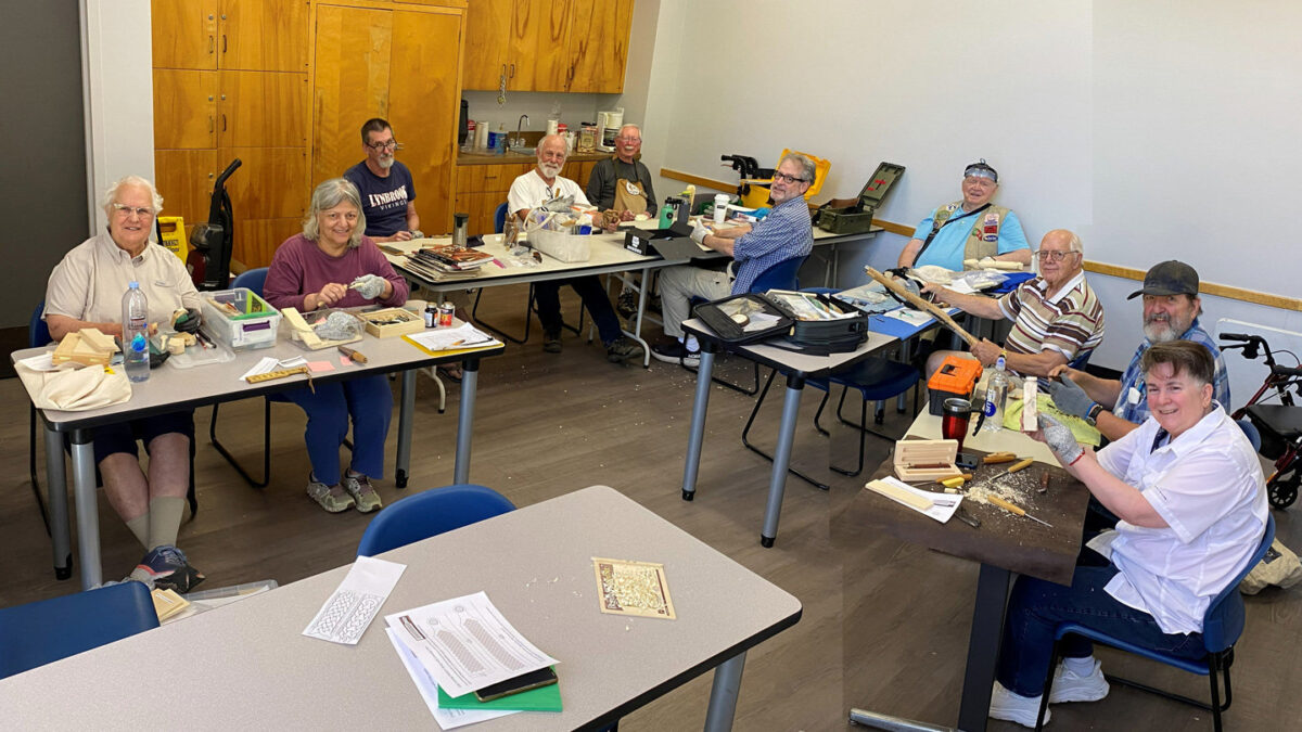 A wide shot captures a gathering of about ten adults seated around tables in a community room, participating in a woodcarving workshop for the Santa Clara Valley Carvers. The participants are surrounded by carving tools, blocks of wood, and shavings. In the foreground on the right, a woman in a white shirt smiles, holding up a carved wooden piece, while sitting next to a bearded man wearing a cap who is working on a driftwood project. On the left, women in beige and purple shirts are seated at a table cluttered with supplies. Along the back wall near the wooden cabinets, several other members are seated, looking towards the camera. The photo was taken for the SCVC woodcarving newsletter.
