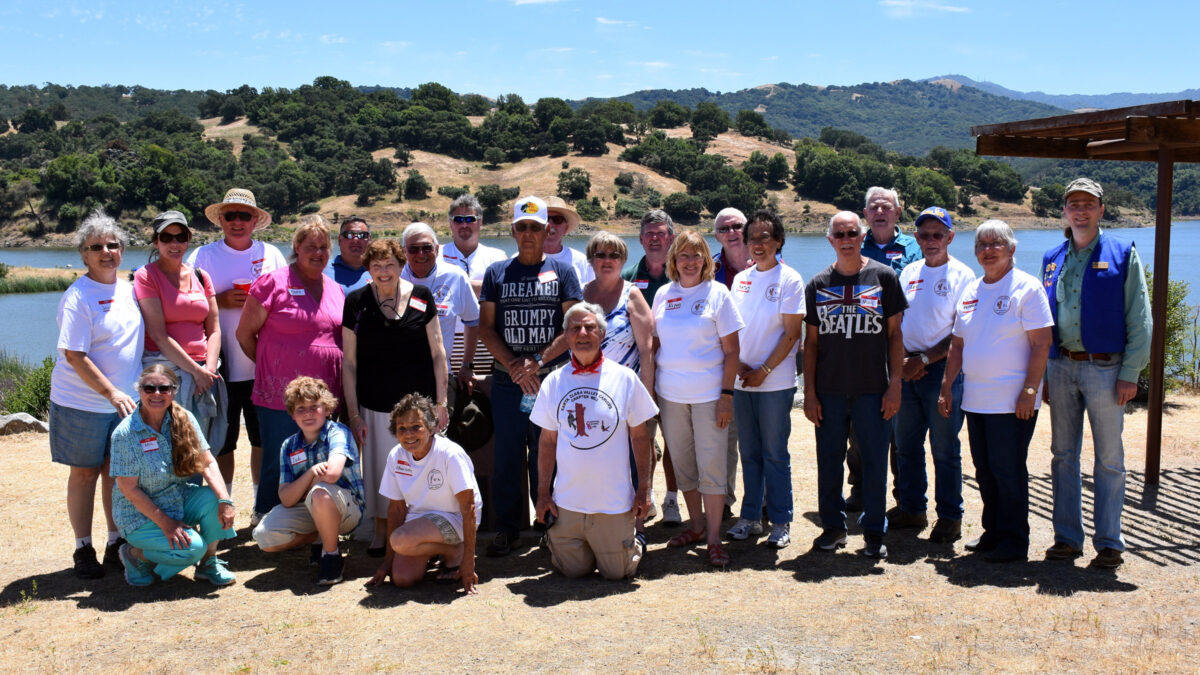 Group photo of about twenty Santa Clara Valley Carvers members and guests at their 2017 annual picnic at Calero Reservoir. Attendees are wearing casual summer attire and club logo T-shirts, set against a backdrop of water and tree-covered hills.