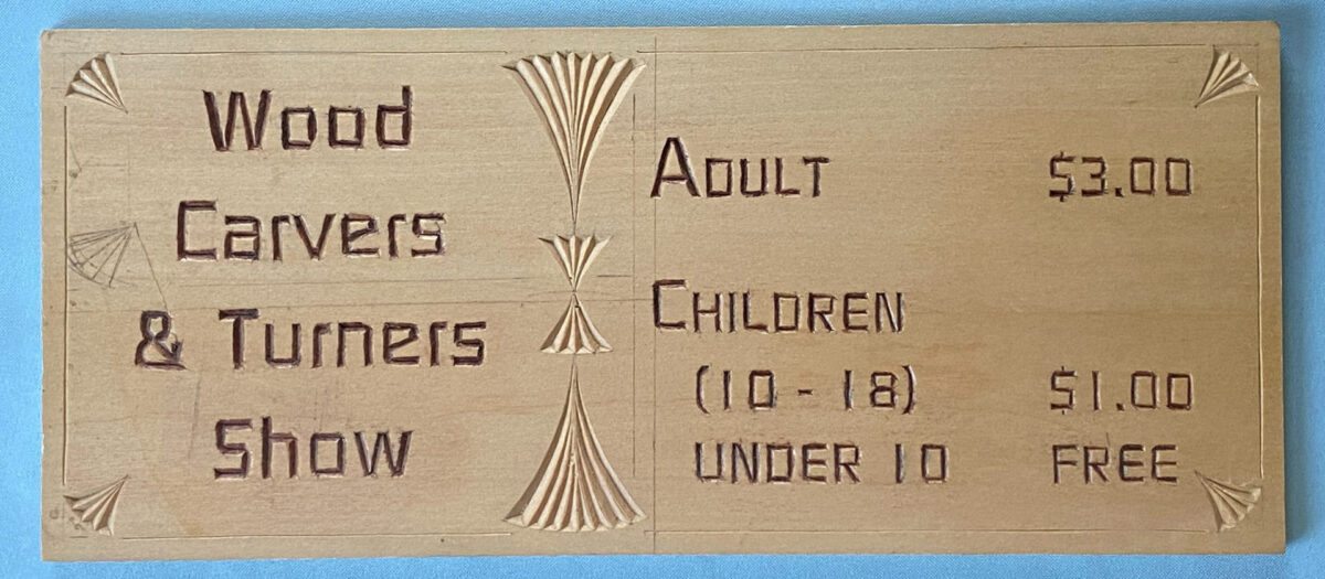 A rectangular wooden sign featuring dark, chip-carved lettering. On the left side, the text reads "Wood Carvers & Turners Show" in stacked block letters. A vertical column of decorative, fan-shaped wood carvings runs down the center, separating the title from the admission prices on the right. The pricing list reads: "ADULT $3.00," "CHILDREN (10 - 18) $1.00," and "UNDER 10 FREE." Small carved fan motifs are visible in each of the four corners.