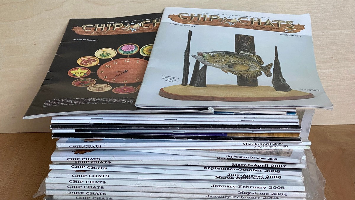A stack of archived Chip Chats woodcarving magazines donated to Santa Clara Valley Carvers by Tri-Valley Carvers rests on a light wooden table.