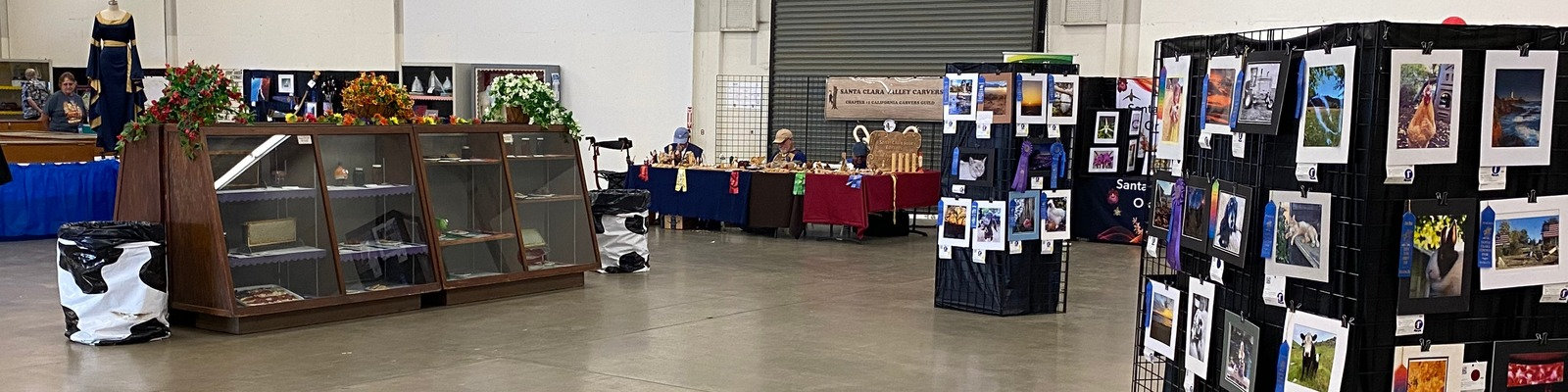 Santa Clara Valley Carvers demonstrate whittling and woodcarving at the Santa Clara County Fair in San Jose. The exhibition booth features award-winning photo displays and members working at red and blue tables.
