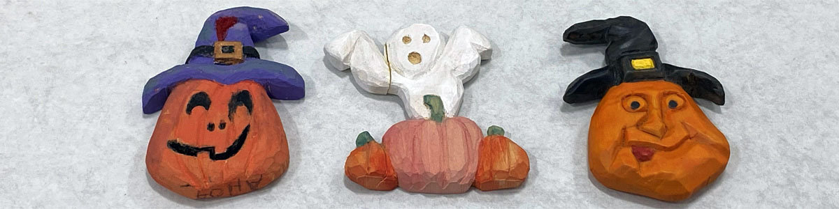 Handcrafted wooden Halloween carvings by Santa Clara Valley Carvers, featuring a jack-o'-lantern in a purple hat, a white ghost, and a witch's head in a black hat.