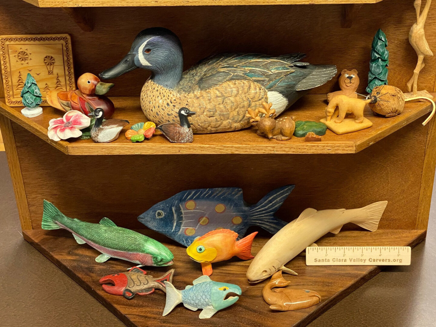 Hand-carved wooden animals and fish in a "Lake View" display by Santa Clara Valley Carvers. The top shelf shows shore animals, and the bottom shelf features various colorful fish carvings.